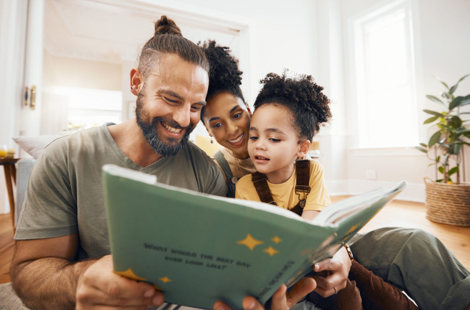 Interracial parents, child and reading with book, story and learning with care, love and teaching on floor. Father, mom and kid with education, knowledge and listening for development in family home A family of three sits on the floor, smiling and reading a book titled "What Would the Next Day Ever Look Like?" in a brightly lit room.