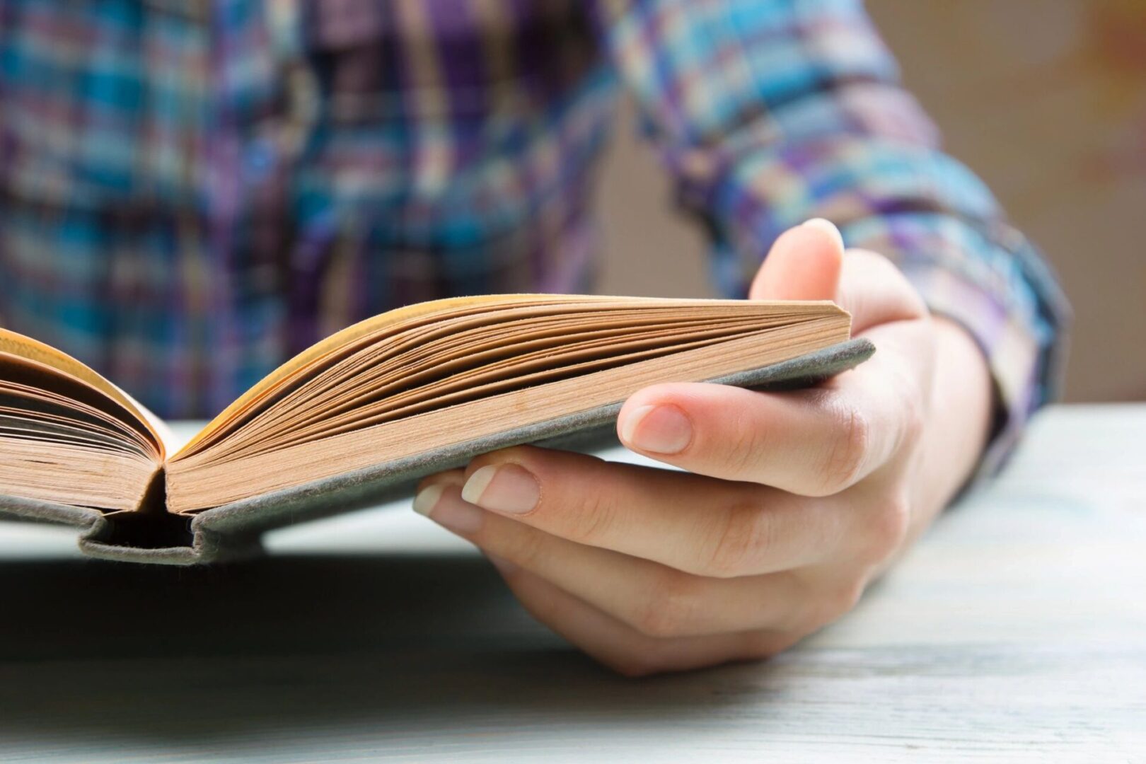 fm=f_21205 A person in a plaid shirt is holding and reading an open book, viewed from the side against a blurred background.