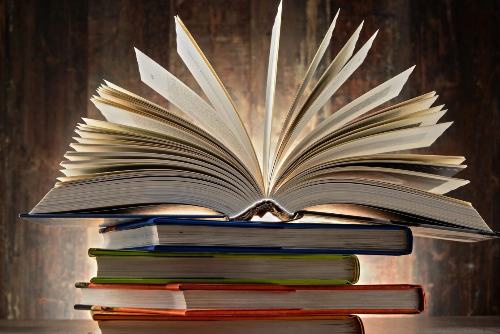 fm=f_28322 An open book with pages fanned out is stacked on top of several closed books against a wooden background.