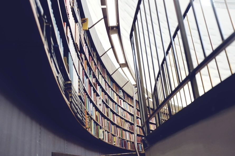 fm=f_99618 Curved library shelves filled with books, viewed from below. A metal railing runs along the right side, beneath a large window.