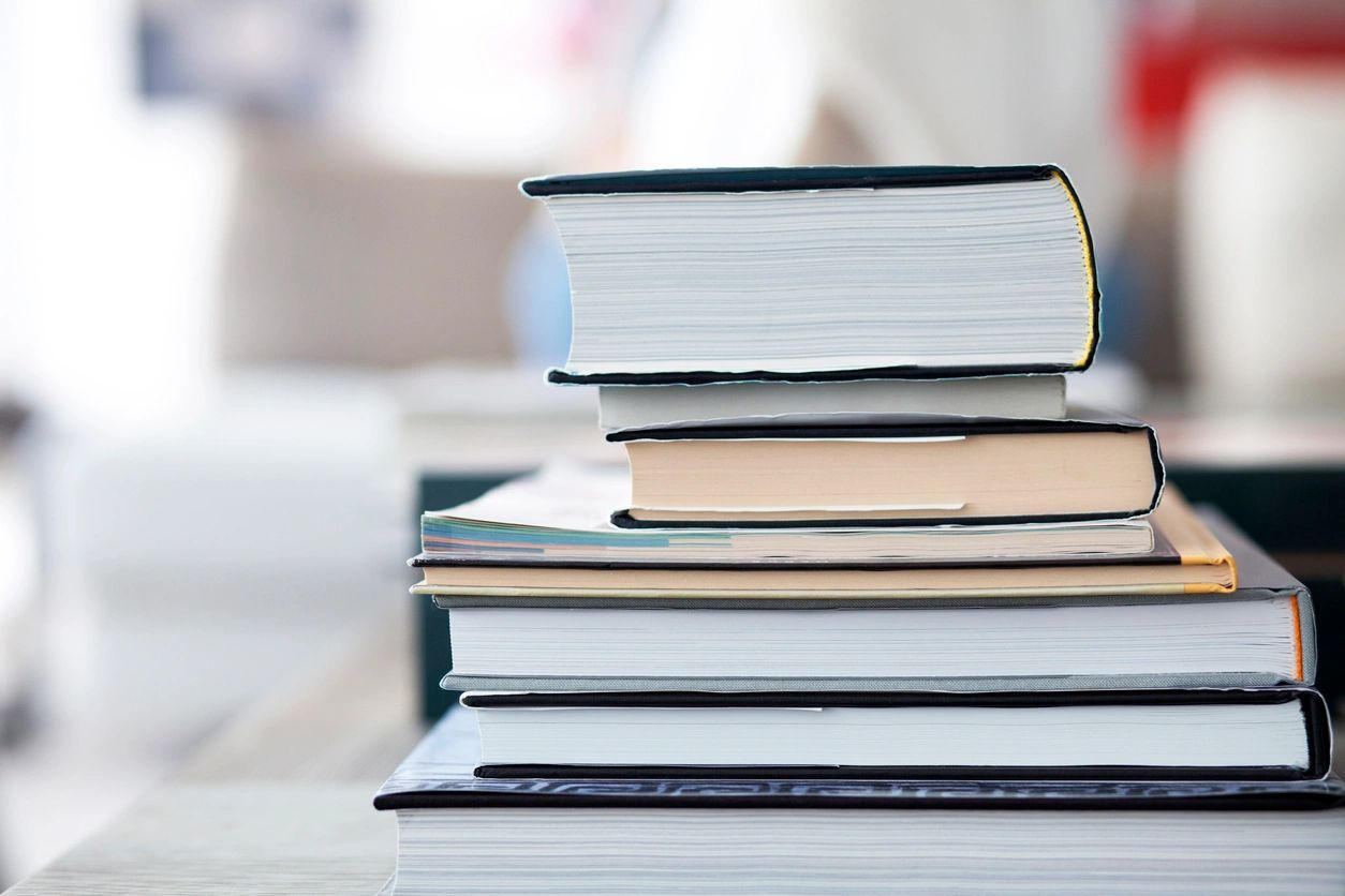 fm=f_opmjVGY A stack of five books lies on a flat surface. The books are closed and arranged unevenly, with various cover colors visible. The background is blurred and indistinct.
