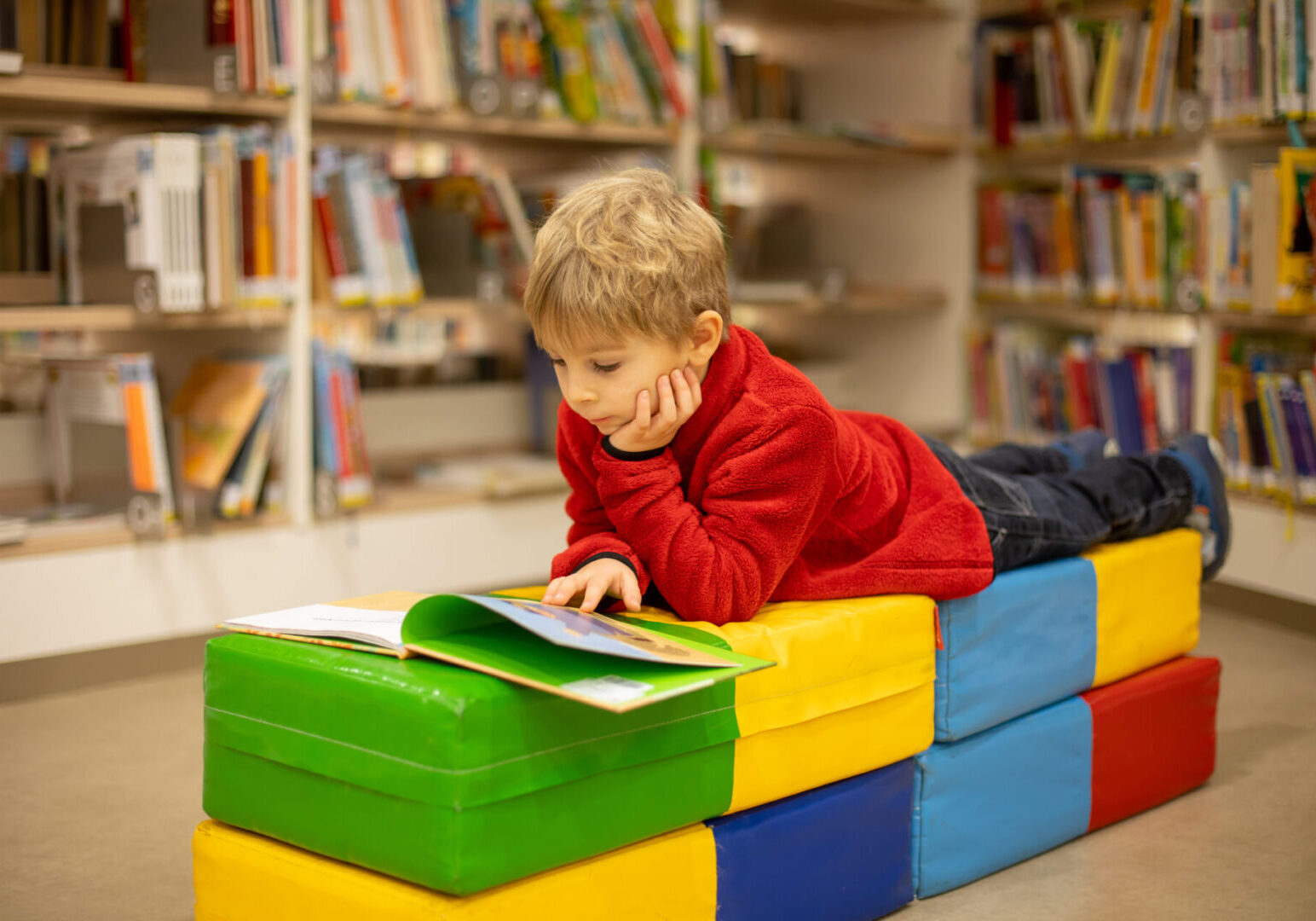 Adorable little boy, sitting in library, reading book and choosing what to lend, kid in book store A child in a red sweater lies on colorful cushions in a library, intently reading a book surrounded by shelves filled with books.
