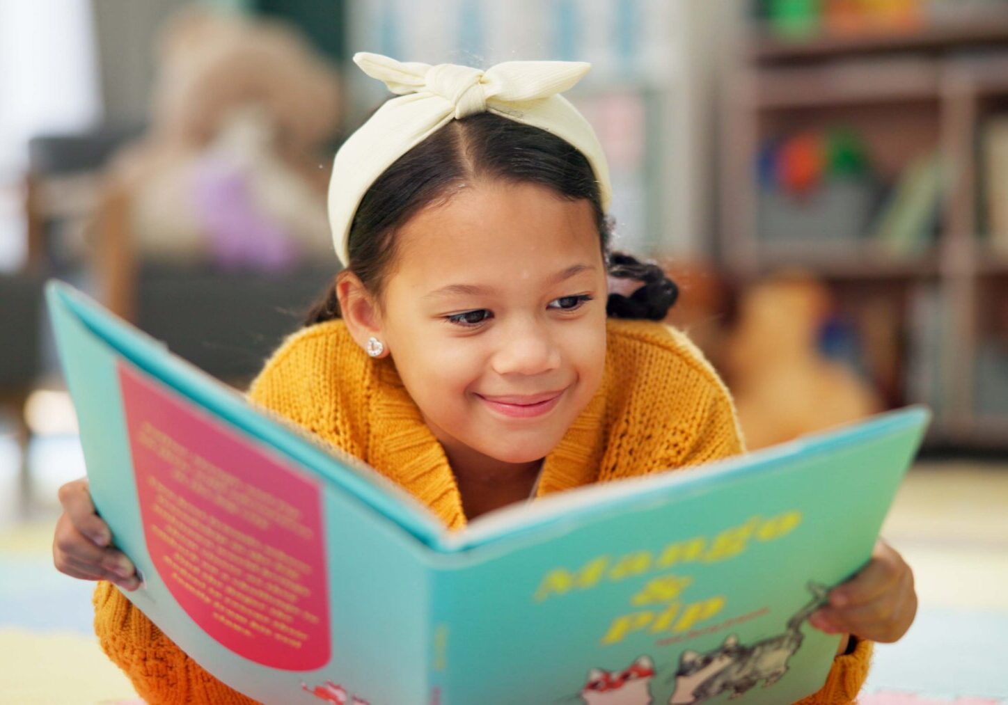 Girl, school and reading book in class, childhood development and growth for vocabulary in kindergarten. Happy, smile and knowledge or information with storybook, learning and fantasy for child Young girl lying on the floor, wearing a white headband and orange sweater, reading a colorful book in a cozy room with bookshelves and toys.
