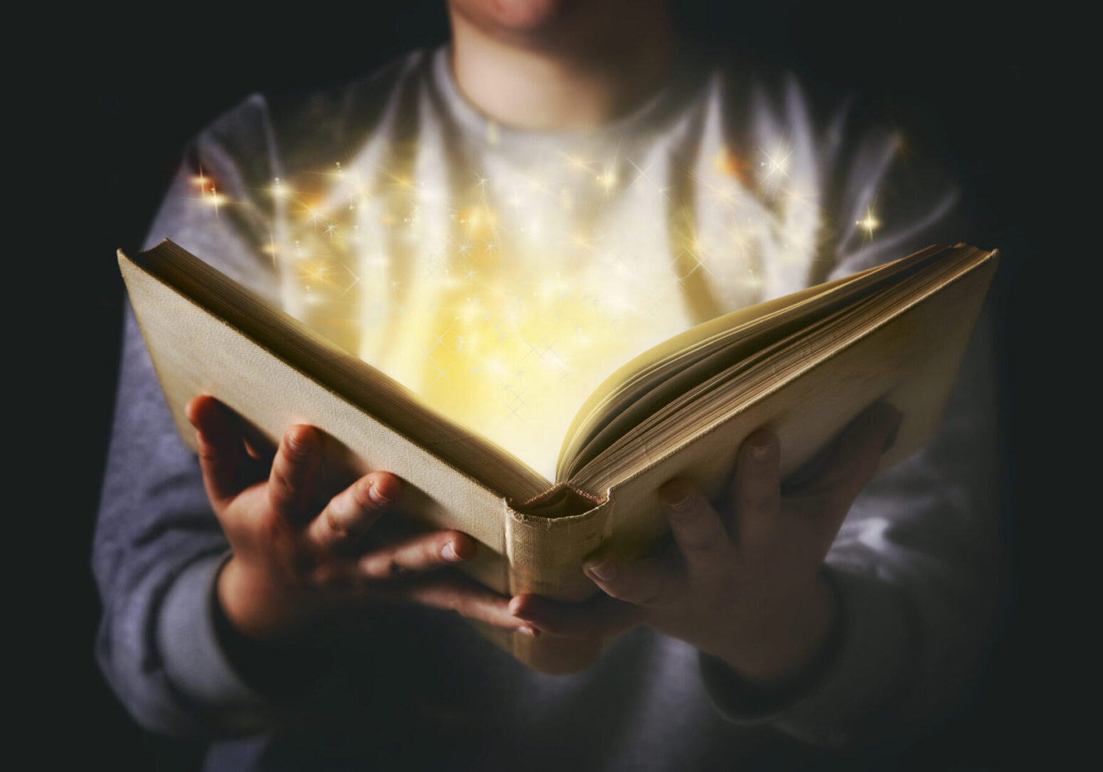 Light coming from book in woman's hands in gesture of giving A person holds an open book emitting glowing golden light and sparkles in a dark setting.