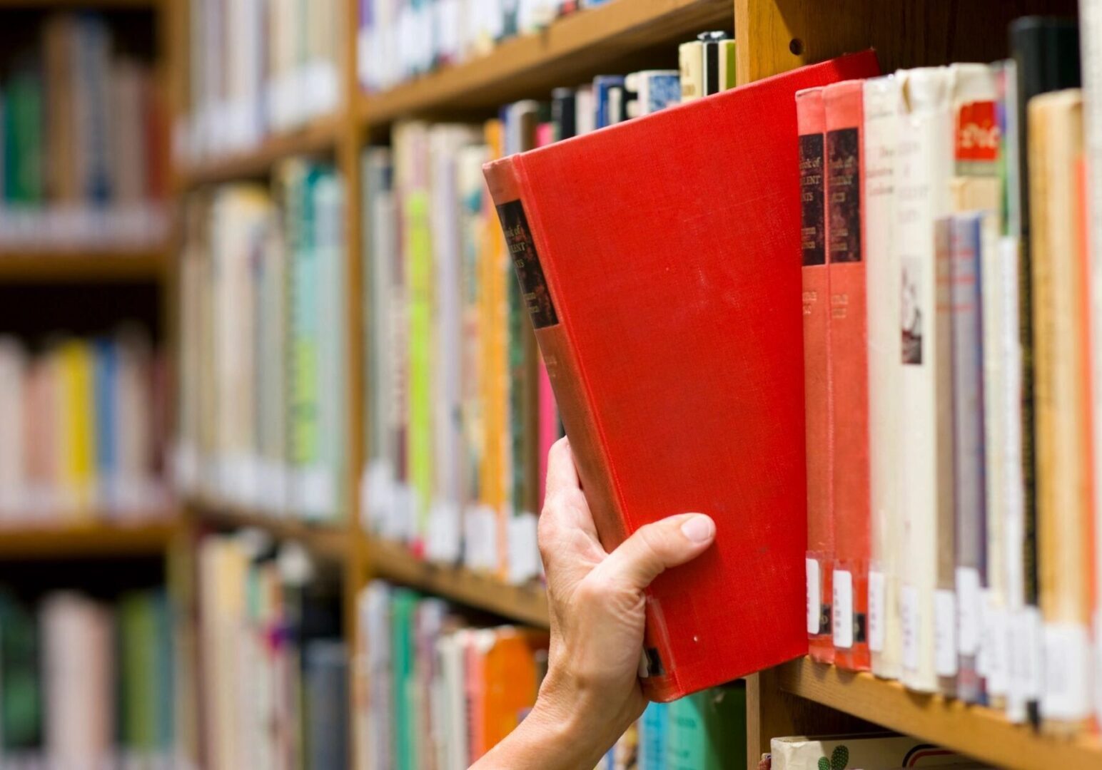 A hand pulls a red book from a packed wooden bookshelf in a library.