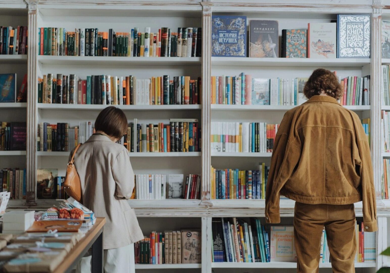 fm=f_upmABV24EmiBBY9zR Two people stand in front of a well-stocked bookshelf in a bookstore, browsing through various books.