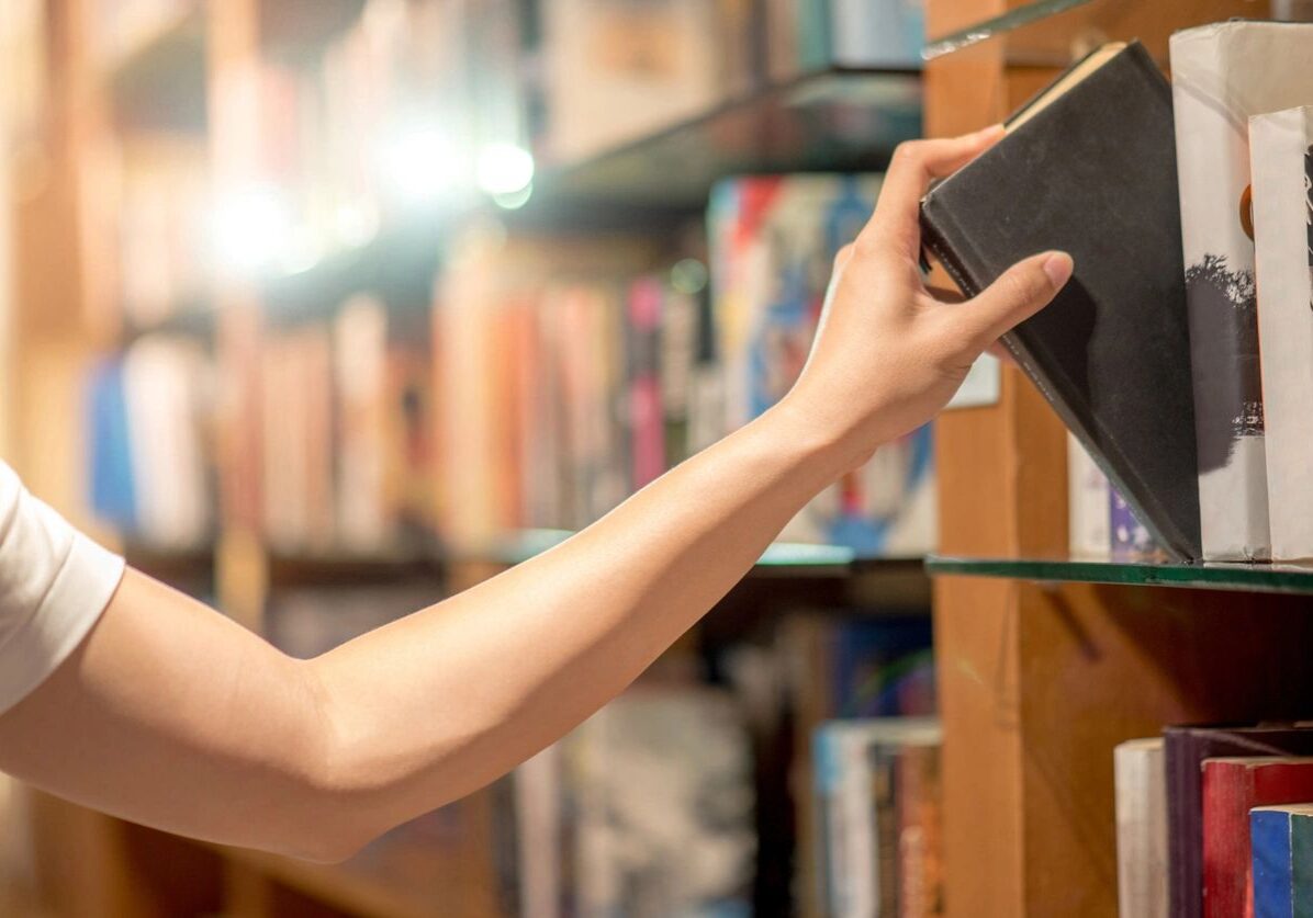 fm=f_yRbOlP0 A person reaches for a black book on a bookshelf in a library or bookstore.
